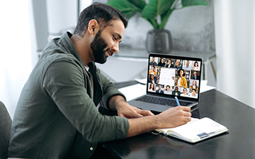 Side view of a successful smart guy listening to an online lecture, taking notes in a notebook, on a laptop screen, a teacher and a group of multiracial people. Online training, webinar
