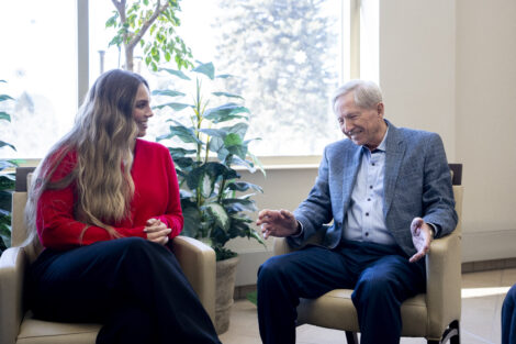 A young woman and an older man sit in armchairs, smiling and engaged in a friendly conversation in a bright room with large windows and green plants.
