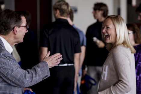 A man and a woman are smiling and laughing while having a lively conversation at a social event, with other people mingling in the background.