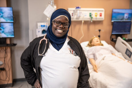 A smiling nursing student wearing a hijab and scrubs standing in a medical simulation lab with a mannequin patient in a hospital bed behind her.