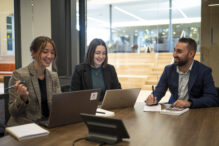 Two women and one man huddled around office table with laptops and notebooks